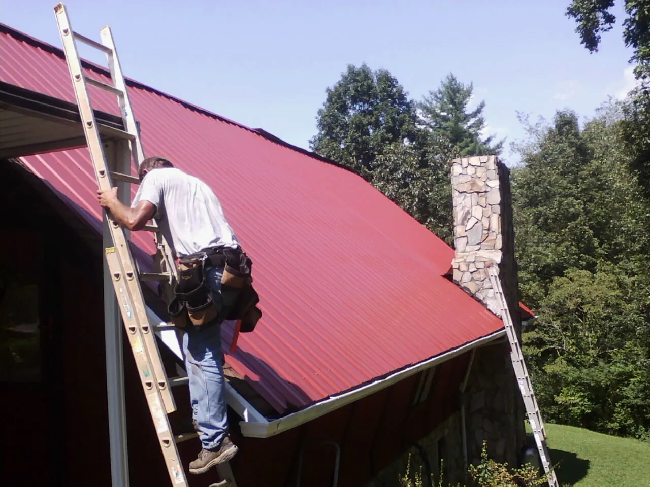 Roofer on a ladder beside a red metal roof and stone chimney — Soffit & Fascia Repair by Roofing Brothers in Pittsburgh